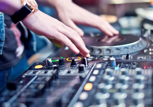 The image shows a DJ's hands operating mixing equipment, with a turntable and various controls visible. The person is wearing a watch.