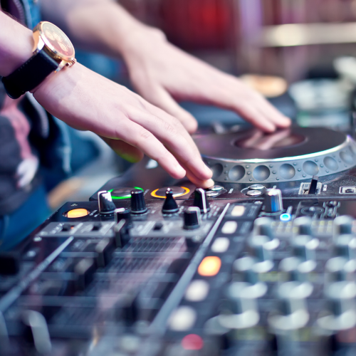 The image shows a DJ's hands operating mixing equipment, with a turntable and various controls visible. The person is wearing a watch.