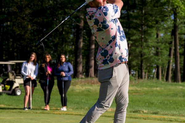 A golfer swings a club on a green, with three spectators and a golf cart in the background, amidst a backdrop of trees.