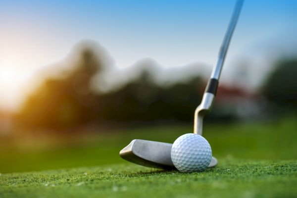 A close-up of a golf club and ball on a green, with a blurry background and a sunset in the distance.