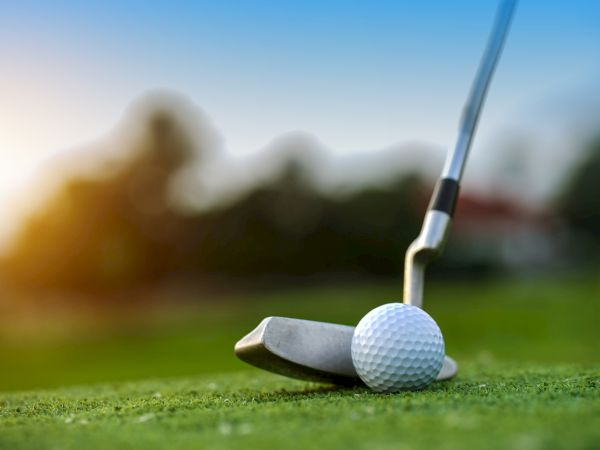 A close-up of a golf club and ball on a green, with a blurry background and a sunset in the distance.