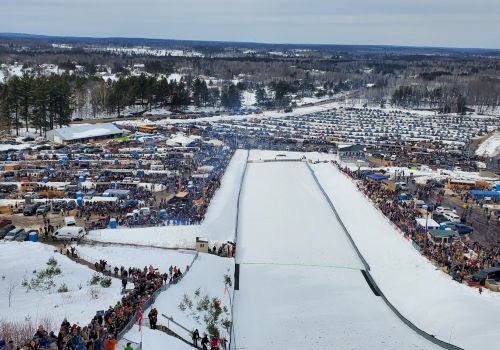 An aerial view of a large outdoor winter event with a snow-covered ramp, numerous spectators, cars, and tents in the background in a forested area.