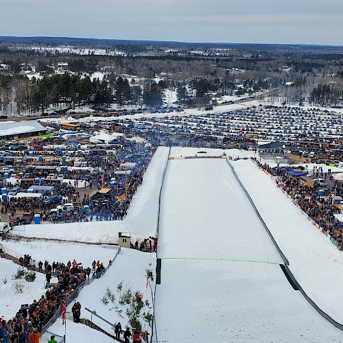 An aerial view of a large outdoor winter event with a snow-covered ramp, numerous spectators, cars, and tents in the background in a forested area.