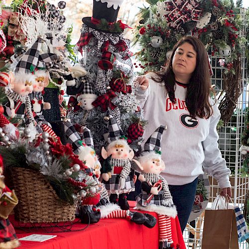 A woman in a white sweater with a shopping bag admires various holiday decorations, including elves and a snowman, displayed on a festive table.