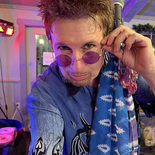 A person wearing purple sunglasses and a blue patterned shirt looks at the camera, holding a microphone. The setting appears to be a casual indoor venue.