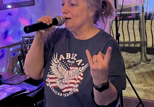 A person is singing into a microphone, wearing a dark T-shirt with an eagle graphic. They are making a hand gesture and appear to be in a casual setting.