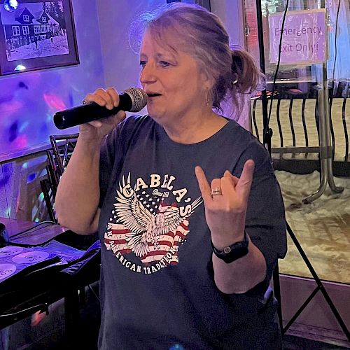 A person is singing into a microphone, wearing a dark T-shirt with an eagle graphic. They are making a hand gesture and appear to be in a casual setting.