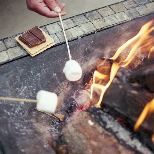 People are roasting marshmallows over a fire pit, preparing ingredients for s'mores with chocolate pieces and graham crackers nearby.