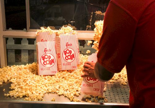 A person filling striped bags with popcorn at a counter, with popcorn scattered around. The bags are labeled "Fresh Popcorn."