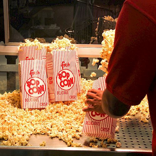 A person filling striped bags with popcorn at a counter, with popcorn scattered around. The bags are labeled 