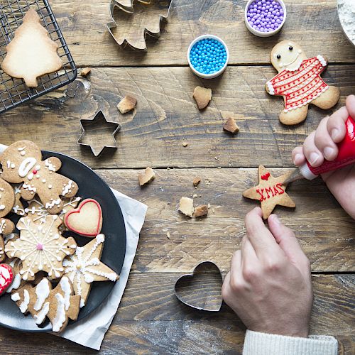 A person decorates a star-shaped cookie with icing. Various Christmas-themed cookies and baking supplies are scattered on a wooden table.