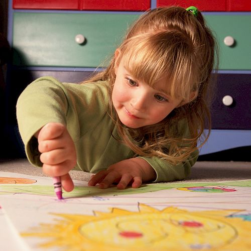 A child is lying on the floor, drawing with crayons on a large sheet of paper, with a colorful drawer set in the background.