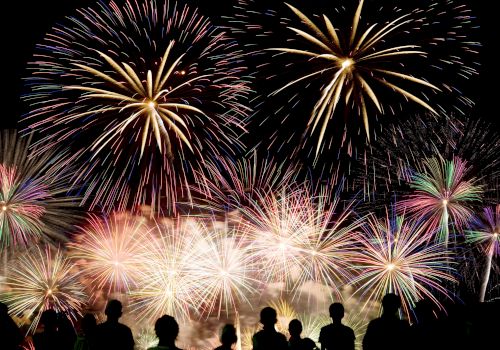 The image shows a night sky lit up with vibrant fireworks in various colors, with a crowd of people silhouetted in the foreground, watching the display.