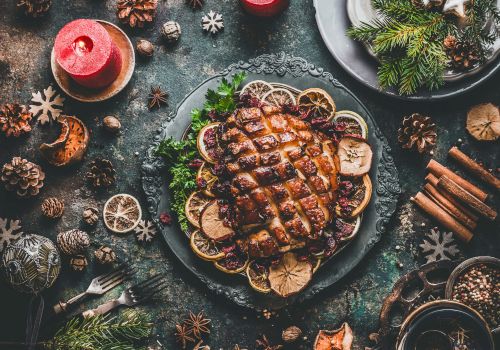 A festive dinner table with a roasted meat centerpiece, candles, spices, pinecones, and rustic holiday decorations surrounding the main dish.