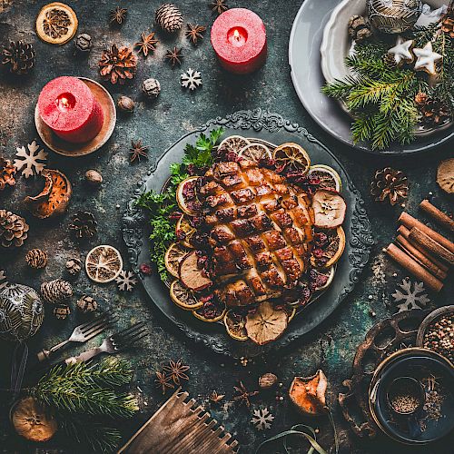 A festive dinner table with a roasted meat centerpiece, candles, spices, pinecones, and rustic holiday decorations surrounding the main dish.