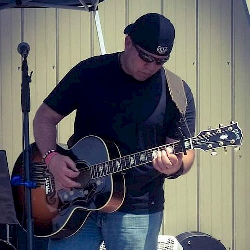 A person wearing a black cap and t-shirt is playing an acoustic guitar on an outdoor stage beside a microphone, with a background of a metallic wall.