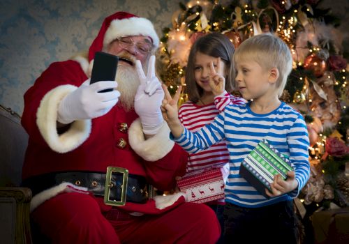 A person dressed as Santa Claus takes a selfie with two children making peace signs, in front of a decorated Christmas tree with lights and ornaments.