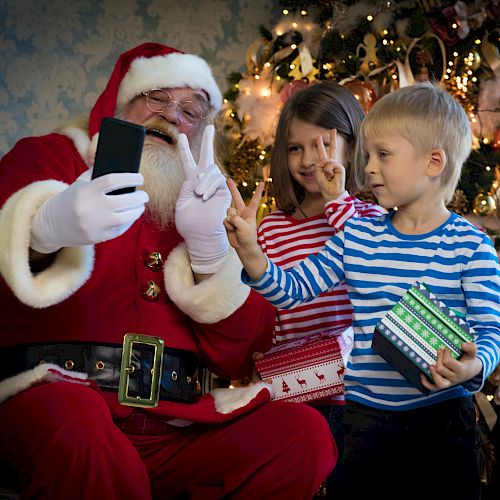 A person dressed as Santa Claus takes a selfie with two children making peace signs, in front of a decorated Christmas tree with lights and ornaments.