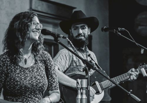 A grayscale image of two musicians performing on stage; a woman is singing, and a man is playing the guitar while wearing a cowboy hat.