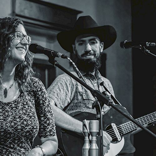 A grayscale image of two musicians performing on stage; a woman is singing, and a man is playing the guitar while wearing a cowboy hat.