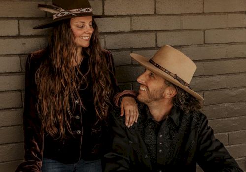 A man and a woman wearing hats are smiling at each other against a brick wall; the man is sitting, and the woman is standing, touching his shoulder.