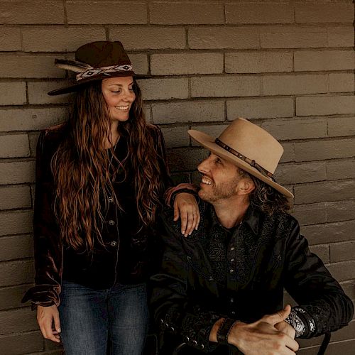 A man and a woman wearing hats are smiling at each other against a brick wall; the man is sitting, and the woman is standing, touching his shoulder.