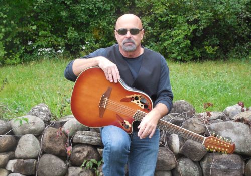 A man wearing sunglasses is sitting on a stone wall, holding an acoustic guitar in an outdoor setting, surrounded by greenery.