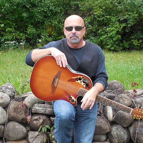 A man wearing sunglasses is sitting on a stone wall, holding an acoustic guitar in an outdoor setting, surrounded by greenery.