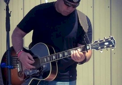 A person dressed in casual clothing plays an acoustic guitar on an outdoor stage, with a microphone stand in the background.