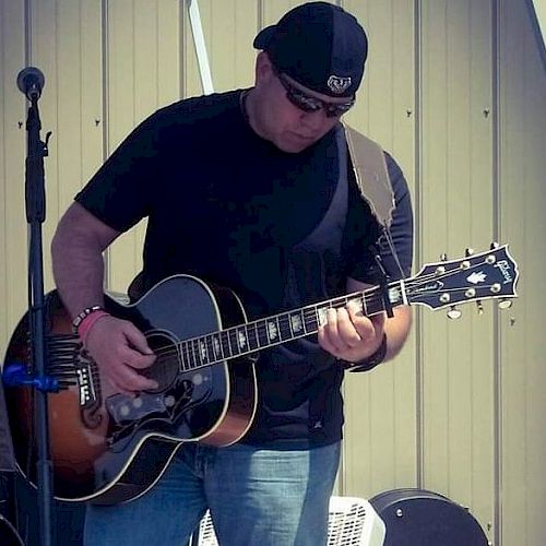 A person dressed in casual clothing plays an acoustic guitar on an outdoor stage, with a microphone stand in the background.