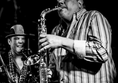 Two men are performing on stage, one playing the saxophone while the other person appears to be cheerfully watching.