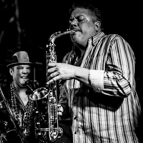 Two men are performing on stage, one playing the saxophone while the other person appears to be cheerfully watching.