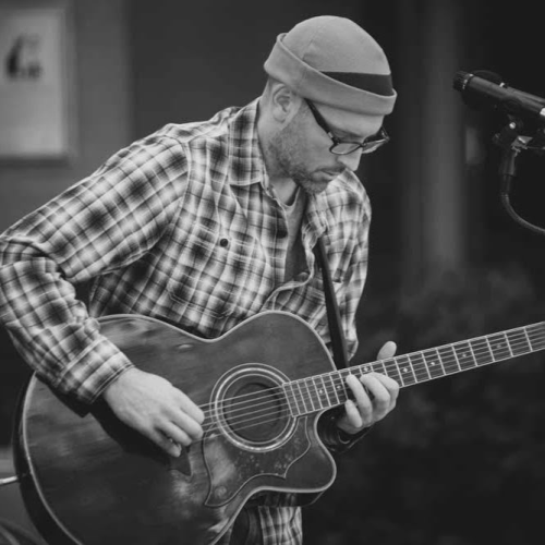 A bearded individual wearing glasses and a beanie plays an acoustic guitar while standing near a microphone, captured in a black-and-white photo.