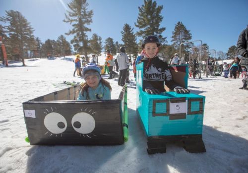 Two children are sitting in snow sleds decorated as cartoon trains, one black and one blue, with number tags.