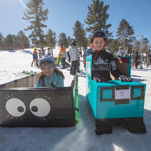 Two children are sitting in snow sleds decorated as cartoon trains, one black and one blue, with number tags.