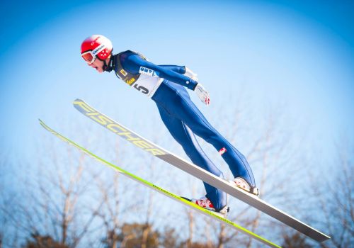 A ski jumper in a blue suit and red helmet is airborne, with Fischer skis, during a clear day.