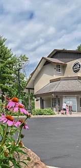 A building with a beige exterior, surrounded by greenery and flowers, with a paved entrance area under a partly cloudy sky.