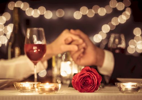 Two people hold hands across a candlelit dinner table, with a red rose and wine glasses. String lights add a romantic ambiance.