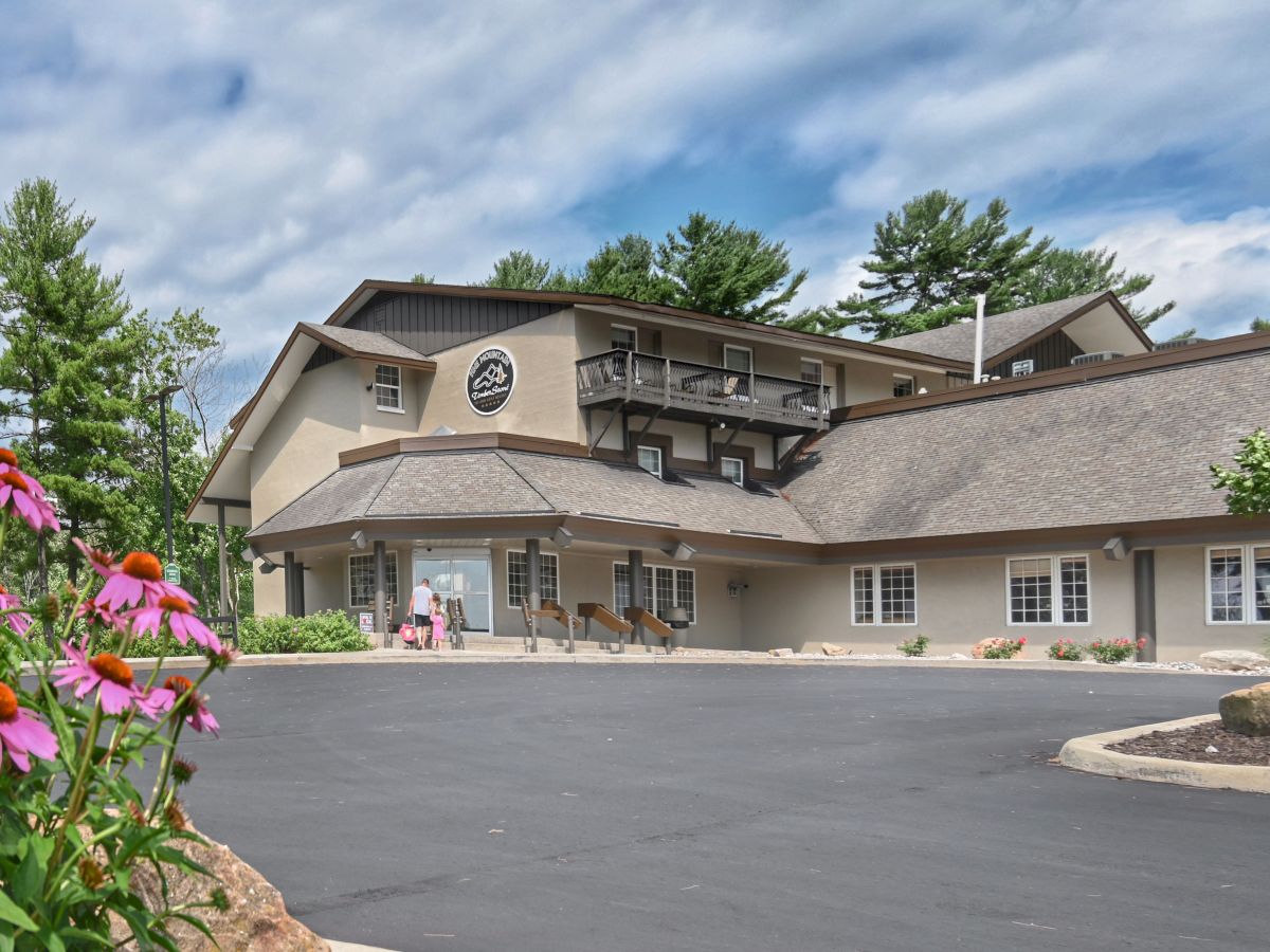 A large lodge-like building with a sloped roof, surrounded by trees and flowers, under a partly cloudy sky, with a spacious parking area in front.