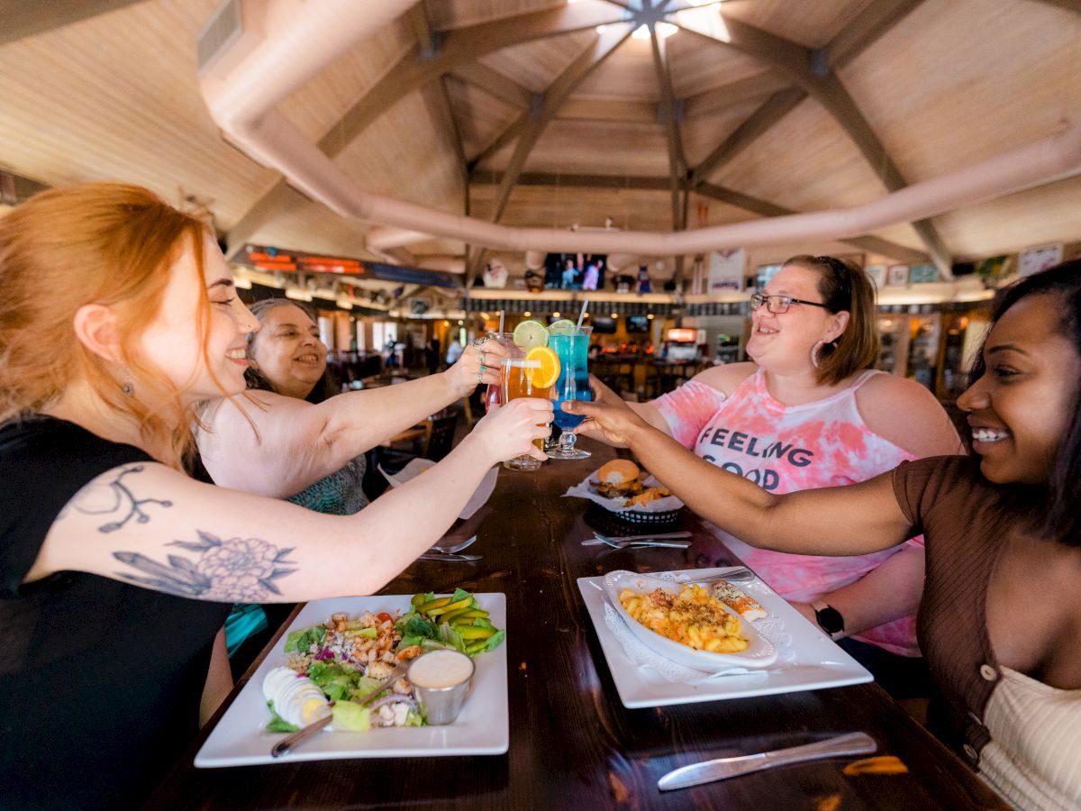 Four people are sitting at a table in a restaurant, toasting with colorful drinks, smiling and enjoying their meal.