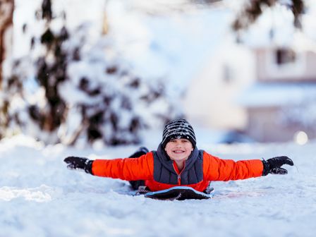 A child in a red jacket and knit hat is lying on a snow-covered ground, smiling with arms outstretched.