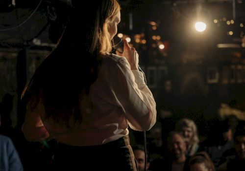 A person on stage is speaking into a microphone, facing an audience, in a dimly lit room with string lights in the background.