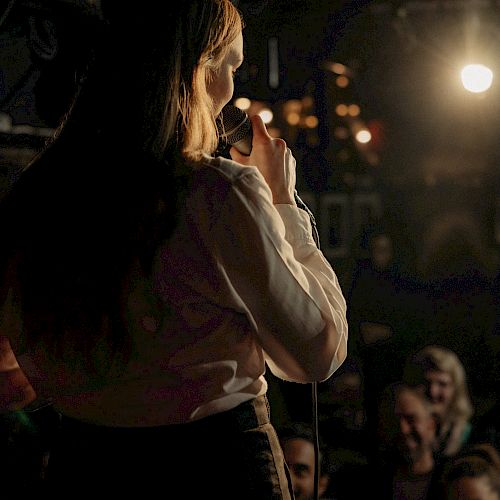 A person on stage is speaking into a microphone, facing an audience, in a dimly lit room with string lights in the background.