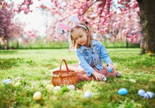 A young girl with bunny ears sits on grass with a basket, surrounded by colorful Easter eggs under blooming trees, enjoying her egg hunt.