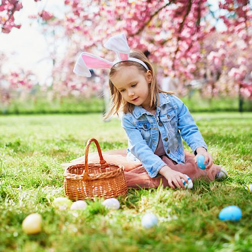 A young girl with bunny ears sits on grass with a basket, surrounded by colorful Easter eggs under blooming trees, enjoying her egg hunt.