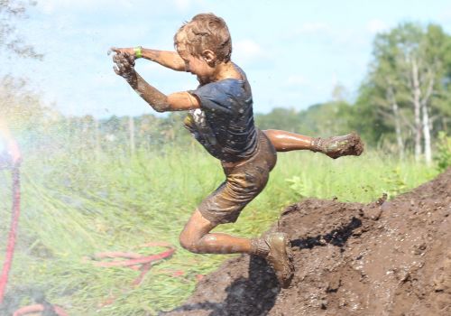 A child, covered in mud, joyfully leaps through the air near a dirt pile, splashing in a muddy field with greenery in the background.