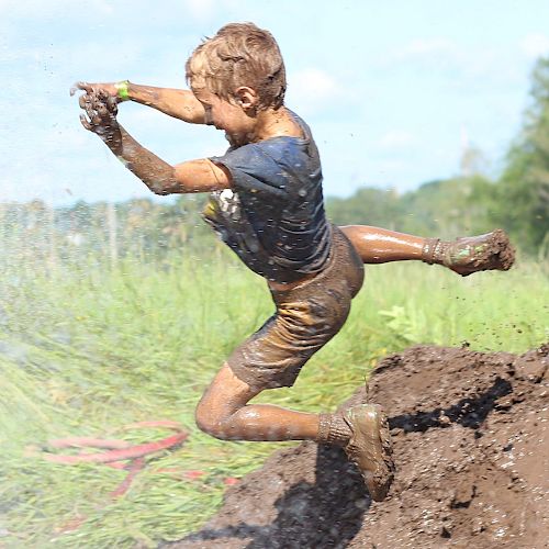 A child, covered in mud, joyfully leaps through the air near a dirt pile, splashing in a muddy field with greenery in the background.