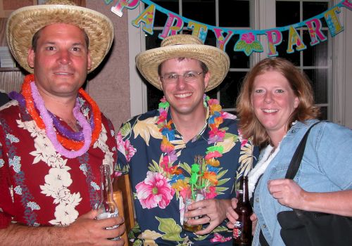 Three people at a party wearing leis and hats, holding drinks. The background has a colorful 