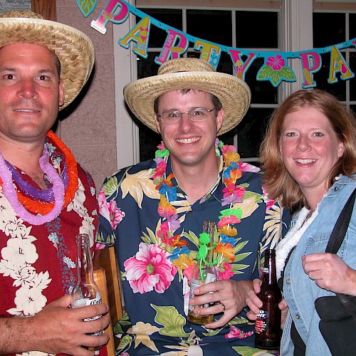 Three people at a party wearing leis and hats, holding drinks. The background has a colorful 