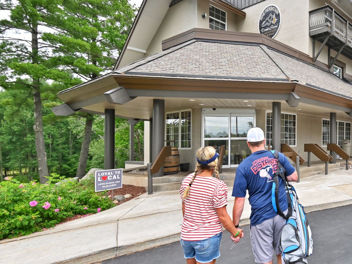 A couple holding hands walks toward a building, with a sign promoting local shopping visible. They're surrounded by trees and greenery.